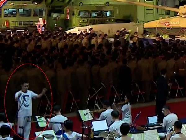 在11日举行的国防发展展览会"自卫-2021"上，指挥演奏歌曲的朝鲜指挥家的T恤（红色圆圈内）上印有朝鲜国务委员长金正恩的头像。在朝鲜，在服装上画最高领导人的头像是非常罕见的情况。因为最高领导人是“神圣不可侵犯的存在”，所以把他的头像印在衣服上很容易被看作是破坏最高领导人的权威的"不良态度"。 朝鲜将印有最高领导人头像的报纸、照片、教科书、书籍等全部规定为"1号出版物"，是在任何情况下都应最先得到照顾和保护的对象。【照片来源：朝鲜中央TV=韩联社】